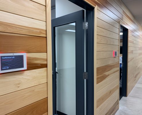 Image shows a wood-panelled hallway leading to CANS Conference Centre. Doors to the facilities are open and digital displays on the wall indicate that the meeting space has been booked.
