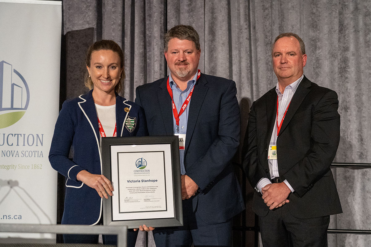 A woman and two men stand on stage smiling. The men are presenting the women with a framed certificate for her service on CANS Board of Directors.