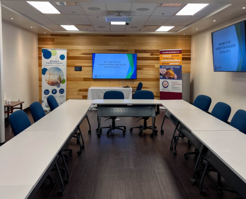 The image shows what CANS Conference space looks like in a u-shape set up. Three boardroom tables are arranged in this shape with CANS Education Banners and an interactive digital display set up in the background.