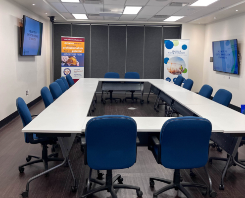 The image shows what CANS Conference space looks like in a u-shape set up. Three boardroom tables are arranged in this shape with CANS Education Banners and an interactive digital display set up in the background. It shows the room from the opposite end from the last photo.