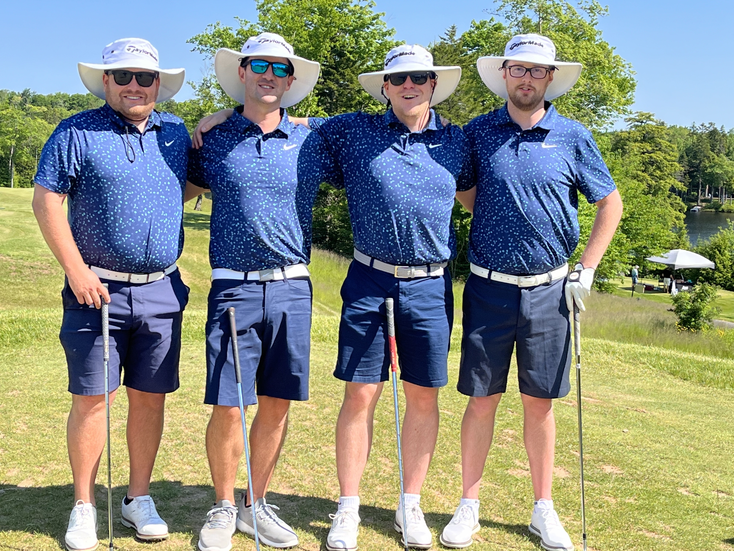 Four male golfers stand in a row posed for a team photo. They are smiling and wearing matching outfits of navy polos, navy, knee-length shorts, white shoes and white Tilley Hats.